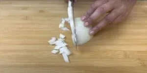 Close-up of hands chopping a white onion on a wooden cutting board.
